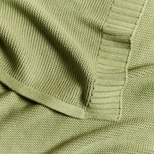 May include: Close-up of a soft, sage green knitted blanket. The blanket has a textured, woven appearance with a ribbed border. The fabric is folded, showing the detail of the knit and the color variations in the light.