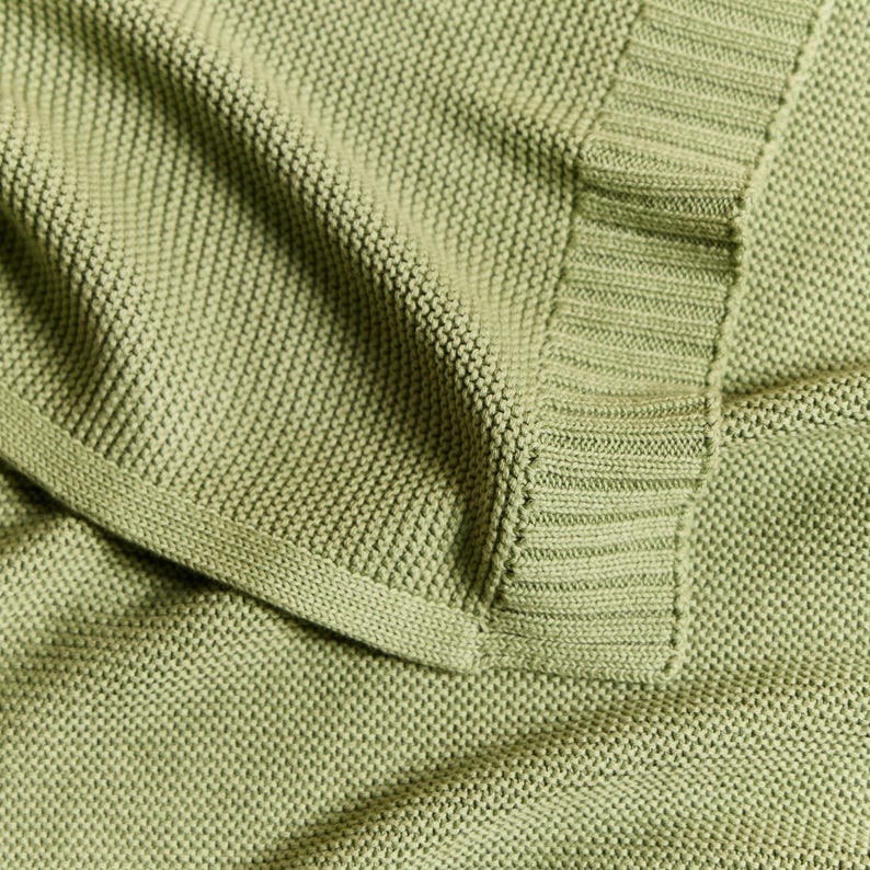 May include: Close-up of a soft, sage green knitted blanket. The blanket has a textured, woven appearance with a ribbed border. The fabric is folded, showing the detail of the knit and the color variations in the light.