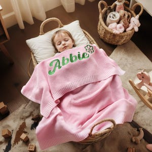 May include: A sleeping child is nestled in a wicker basket, covered with a pink blanket. The blanket is embroidered with the name "Abbie" in green script. A white pillow supports the child's head. A basket of toys sits nearby.