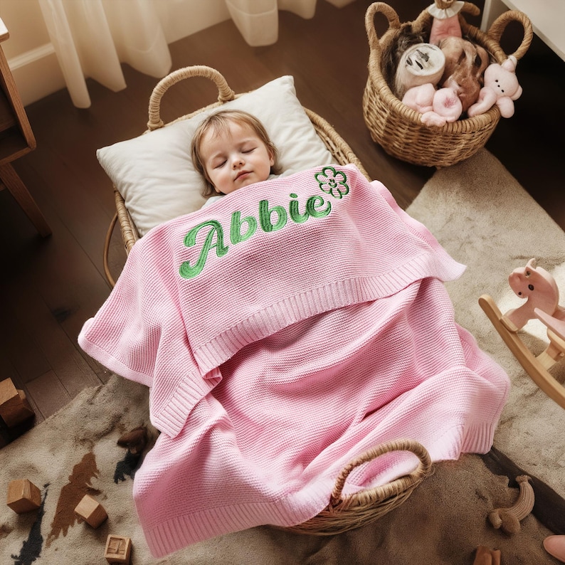 May include: A sleeping child is nestled in a wicker basket, covered with a pink blanket. The blanket is embroidered with the name "Abbie" in green script. A white pillow supports the child's head. A basket of toys sits nearby.