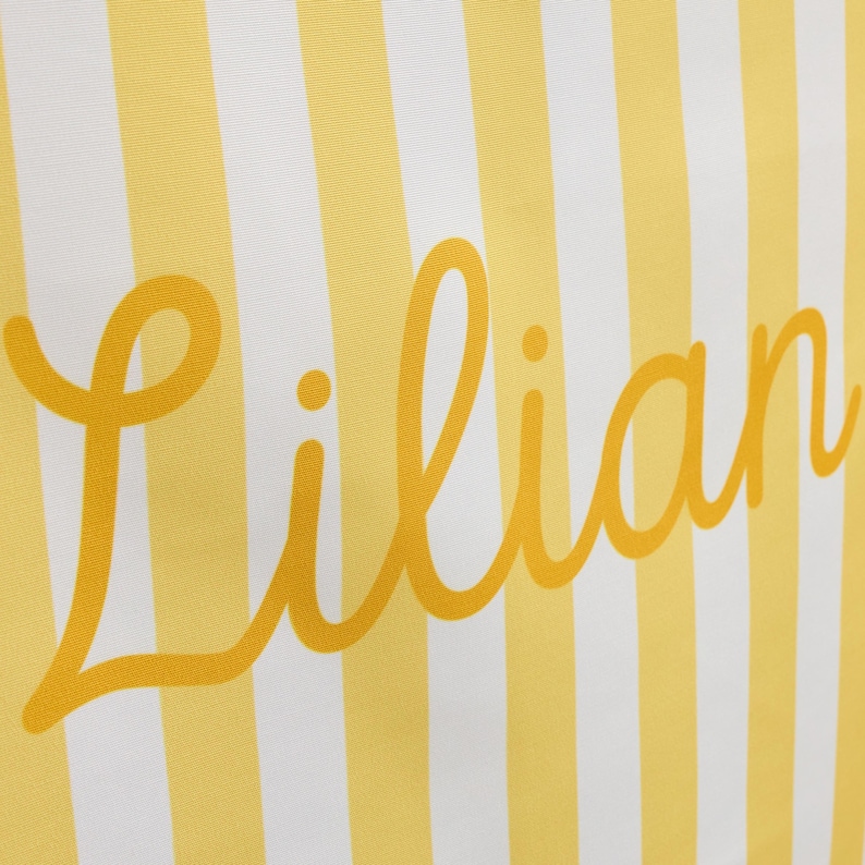 K&ouml;nnte beinhalten: Eine Stoffoberfl&auml;che mit einem Muster aus vertikalen gelben und wei&szlig;en Streifen. Der Name Lilian ist in der Mitte in einer gro&szlig;en, kursiven, gelben Schriftart aufgedruckt. Das Design ist &uuml;ber den gesamten sichtbaren Bereich einheitlich.