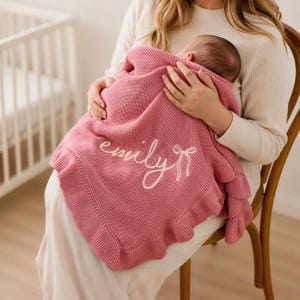 May include: A dusty rose knitted baby blanket featuring a ruffled border and the name emily embroidered in white cursive script, accompanied by a small white embroidered bow detail on the right side of the text, creating a soft texture.
