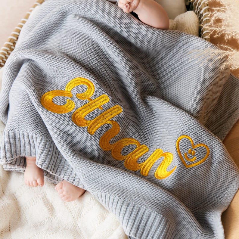 May include: A gray knitted baby blanket with the name "Ethan" embroidered in yellow script. A yellow heart with a smiling face is embroidered next to the name. The blanket is folded over a baby.