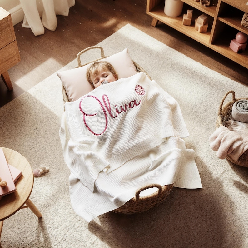 May include: A sleeping baby in a woven basket, nestled on a pillow and covered with a white blanket. The blanket is embroidered with the name "Oliva" in a cursive font and a floral design. The basket is on a light-colored rug.
