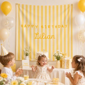 K&ouml;nnte beinhalten: Ein gelb-wei&szlig; gestreifter Stoffhintergrund mit dem Text HAPPY BIRTHDAY und dem Namen Lilian in gelber Schreibschrift. Das Banner befindet sich hinter einem Tisch mit einem Geburtstagskuchen, Blumen und Geschenken, umgeben von Luftballons.