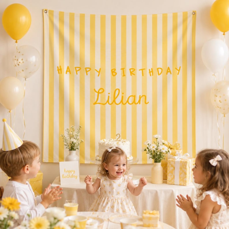 K&ouml;nnte beinhalten: Ein gelb-wei&szlig; gestreifter Stoffhintergrund mit dem Text HAPPY BIRTHDAY und dem Namen Lilian in gelber Schreibschrift. Das Banner befindet sich hinter einem Tisch mit einem Geburtstagskuchen, Blumen und Geschenken, umgeben von Luftballons.
