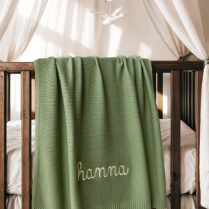 May include: A sage green knitted baby blanket draped over a wooden crib. The blanket features the embroidered name "hanna" in a cursive font. A white mobile with three flying geese hangs above the crib.