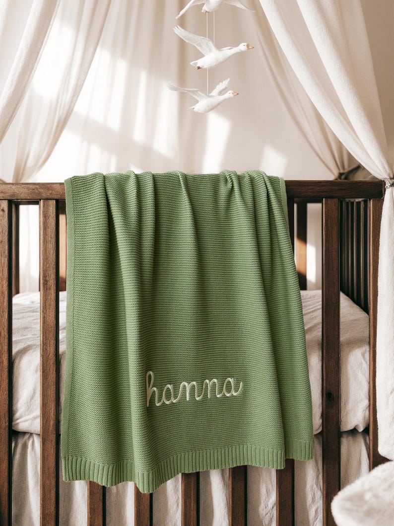 May include: A sage green knitted baby blanket draped over a wooden crib. The blanket features the embroidered name "hanna" in a cursive font. A white mobile with three flying geese hangs above the crib.
