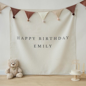 May include: A cream-colored backdrop with the text "HAPPY BIRTHDAY EMILY" is displayed. A garland of triangular flags in shades of brown and cream hangs above. A teddy bear and a small cake with candles are in front.