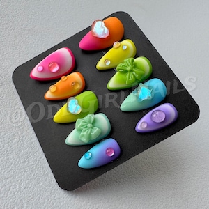 Peut inclure: Une collection d'ongles artificiels en forme d'amande aux couleurs vives. Chaque ongle présente un design unique, avec des dégradés, des nœuds et des ornements en forme de cœur. Les ongles sont disposés sur une surface noire, mettant en valeur une variété de couleurs et de textures.