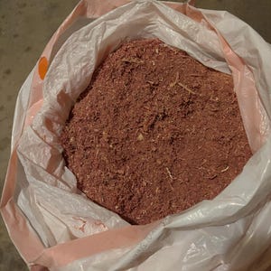 Könnte beinhalten: Ein großer, offener weißer Plastikbeutel gefüllt mit rötlich-braunen Holzspänen. Der Beutel hat eine rosa Umrandung und ist teilweise geöffnet, wodurch die Textur und Farbe des Holzmaterials sichtbar wird. Die Späne scheinen frisch und trocken zu sein.