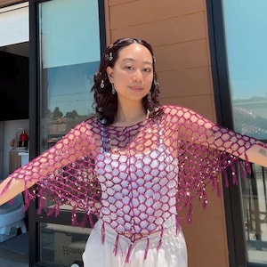 May include: A dark magenta crochet poncho featuring an open net pattern with integrated reflective sequins throughout the fabric. The garment has a wide neckline and is finished with a fringe trim along the bottom edge, creating a textured look.