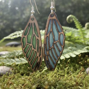 May include: A pair of handcrafted wooden earrings shaped like butterfly wings. One earring features green and brown tones, while the other has blue and brown hues. The earrings hang from silver-colored hooks, set against a natural, mossy background.