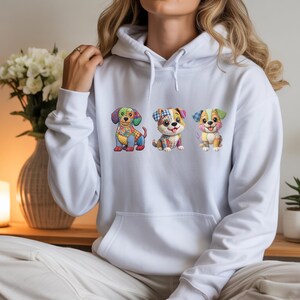 Puede incluir: Una sudadera con capucha blanca que presenta un estampado gráfico de tres perros de dibujos animados. Cada perro está diseñado con un colorido patrón de patchwork compuesto por varias formas geométricas, motivos florales y cuadros en tonos azul, rojo y amarillo.