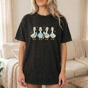 Könnte beinhalten: Schwarzes T-Shirt mit einer Grafik von vier Cartoon-Gänsen in Krankenschwester-Uniformen. Jede Gans trägt eine Krankenschwesterhaube und hält medizinische Geräte. Die Gänse stehen in einer Reihe, mit einer Spritze, einer Arzttasche und einem Stethoskop.