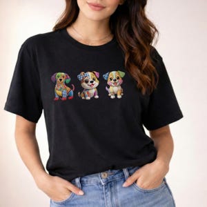 May include: A black short-sleeve t-shirt featuring a graphic print of three small dogs. Each dog is rendered in a colorful patchwork style with various patterns including plaid, floral, and polka dots in shades of blue, green, yellow, and pink.