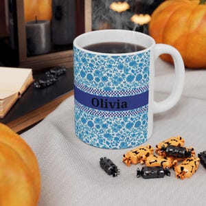 Puede incluir: Taza de cerámica blanca con un estampado floral azul y el nombre "Olivia" en una banda azul marino. La taza contiene un líquido oscuro y está sobre una superficie gris con caramelos de Halloween.