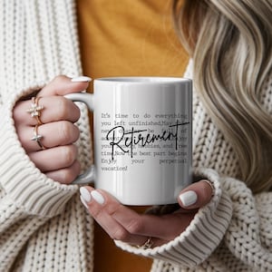 Puede incluir: Taza de cerámica blanca con la palabra "Jubilación" en letra cursiva. La taza tiene un asa gris y presenta texto sobre disfrutar de la jubilación y las vacaciones. La taza la sostiene una persona que lleva un suéter color crema.