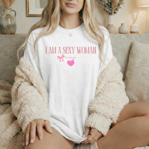 May include: White t-shirt with the text "I AM A SEXY WOMAN at my age" in pink, along with a pink bow and heart graphic. The shirt is a casual style, suitable for everyday wear.