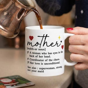 Puede incluir: Taza de cerámica blanca con la palabra "mother" en escritura negra, adornada con corazones rojos y una estrella amarilla. La taza incluye una definición y frases de apoyo. Se está vertiendo café en la taza.