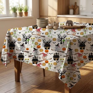 May include: A white tablecloth featuring a repeating Halloween pattern of cartoon mummies, cats in witch hats on broomsticks, elephants in witch hats, black haunted houses, orange pumpkins, and small black bats scattered across the fabric.