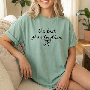 Puede incluir: Camiseta verde claro con la frase "the best grandmother" en escritura negra y un lazo. La camiseta tiene mangas enrolladas y un corte holgado. Un estilo cómodo e informal.