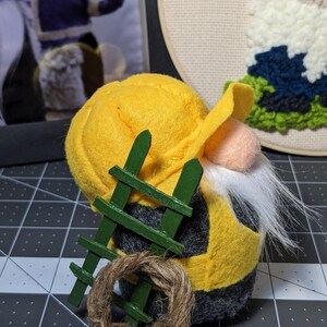 May include: A handmade gnome with a yellow hat and vest, gray body, and white beard. It has a small green ladder and a twine wreath. The gnome is a decorative item.