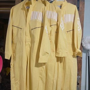 Puede incluir: Tres monos de trabajo amarillos colgados en un perchero. Cada mono tiene cierre de cremallera, múltiples bolsillos y un diseño de código de barras blanco en el pecho. Un mono tiene un diseño tribal blanco en la manga.