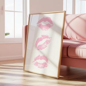 Può includere: Stampa artistica incorniciata con tre impronte di baci di rossetto rosa su sfondo crema. L'opera d'arte è presentata in una cornice dorata e appoggiata a un divano rosa. L'immagine è ambientata in una stanza ben illuminata con una finestra.
