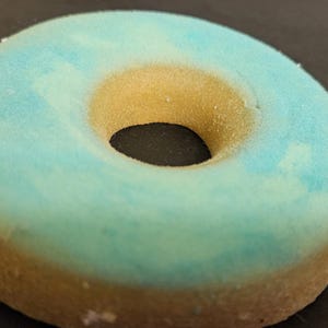 May include: A close-up shot of a frosted donut. The donut has a light blue frosting on top and a golden-brown base. The hole in the center is dark. The donut is sitting on a dark surface.