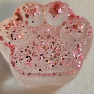 Könnte beinhalten: Ein pfotenförmiger Gegenstand aus Harz mit klarer Basis, großzügig mit rosa und roségoldenen Glitzer bestreut. Der Glitzer verleiht dem Pfotenabdruck-Design Glanz und Dimension und erzeugt einen dekorativen Effekt.