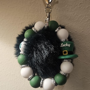 Puede incluir: Un llavero con temática del Día de San Patricio con un centro de pelusa negra, cuentas verdes y blancas y un dije de sombrero verde que dice "Lucky". El llavero tiene un cierre plateado.