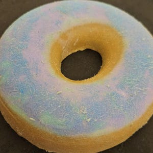 May include: A close-up of a donut-shaped object with a light blue and yellow color scheme. The top surface has a light blue hue with hints of green and purple, while the sides and center are yellow. The object is on a dark surface.