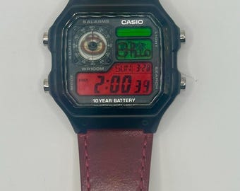 May include: Black Casio digital watch with a burgundy leather strap. The watch face shows the time, date, and other functions. The watch has the text "CASIO" and "10 YEAR BATTERY" printed on the face.