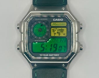 Peut inclure: Une montre numérique Casio argentée avec un écran vert affichant l'heure et la date. La montre a un bracelet en cuir vert et propose plusieurs fonctions, dont des alarmes et l'heure mondiale. Le cadran affiche le texte "CASIO" et "10 YEAR BATTERY".