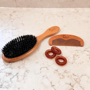 以下が含まれることがあります： 木製のヘアブラシ、櫛、3つの茶色のスパイラルヘアタイが、大理石の表面に配置されています。ブラシと櫛には「Egg」の文字がプリントされています。ブラシには黒い毛が付いています。