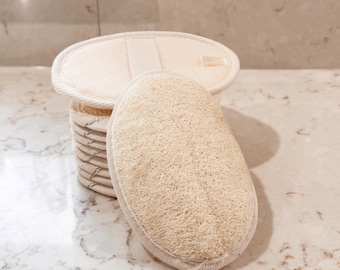 Op de afbeelding: Een stapel beige en witte geweven containers met een luffaspons. De luffa is ovaal van vorm en heeft een lichtbeige kleur. De containers zijn cilindrisch en hebben een geweven ontwerp. De items staan op een marmeren aanrecht.