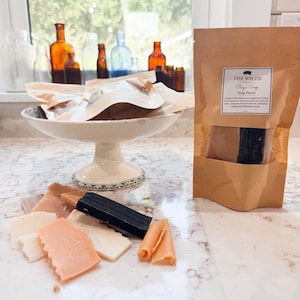 Puede incluir: Una exhibición de piezas de jabón hechas a mano en varios colores y formas. Se ve una bolsa de papel marrón con el texto "THE WYLD'S Big's Soap Pieces". Un cuenco de cerámica blanco contiene más piezas de jabón, con botellas de vidrio antiguas en el fondo.