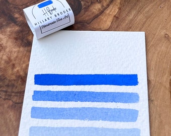 Può includere: Un'opera d'arte ad acquerello con quattro pennellate blu orizzontali di varie tonalità, dal profondo al chiaro. Un piccolo pacchetto con la scritta "Hillary Broder Handmade Fine Art" è accanto all'opera d'arte. Un minerale blu scuro è appoggiato su una superficie di legno.