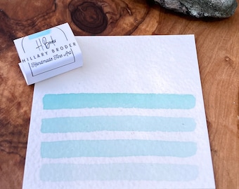 Può includere: Cinque campioni di acquerello di un colore blu chiaro su un cartoncino bianco. Il cartoncino è su una superficie di legno. Una piccola scatola con il testo "Hilary Broder Handmade Fine Art" si trova nell'angolo in alto a sinistra dell'immagine.