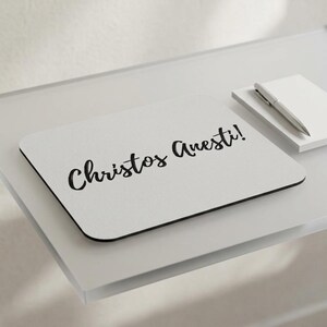 May include: A rectangular mouse pad with a light gray surface featuring the black cursive text Christos Anesti! printed in the center. The mouse pad has rounded corners and a thin black border, positioned next to a white notepad and a silver pen.