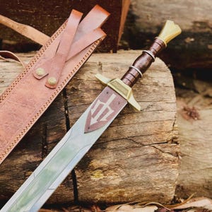 Op de afbeelding: Een decoratief zwaard met een bruin handvat en een zilveren lemmet, rustend op een houten oppervlak. Het zwaard heeft een drietand-embleem en gouden accenten. Een bruine leren schede is zichtbaar op de achtergrond.