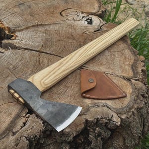 Puede incluir: Un hacha artesanal con mango de madera y cabeza de metal oscuro, apoyada sobre un tocón de árbol. Una funda de cuero marrón con cierre de botón a presión está junto al hacha. La hoja del hacha mide unos 10 cm de ancho.