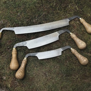 Puede incluir: Tres cuchillos de tracción con hojas curvas y mangos de madera. Las hojas son plateadas y los mangos son de color marrón claro con un diseño de vetas de madera. Las herramientas están dispuestas sobre un lecho de musgo verde.