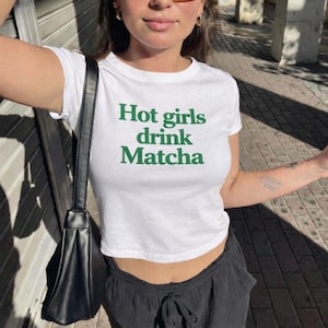 May include: White crop top with the text "Hot girls drink Matcha" in green. The shirt has short sleeves and a crew neck. The model is wearing black pants and a black shoulder bag.
