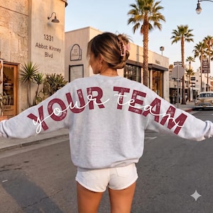 Könnte beinhalten: Ein hellgraues Sweatshirt mit Rundhalsausschnitt und dem Schriftzug YOUR TEAM auf der Rückseite in einer abgenutzten, dunkelroten Blockschrift, überlagert von einer weißen Schreibschrift. Das Sweatshirt hat lange Ärmel und eine lockere Passform.