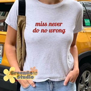 May include: A light gray t-shirt with the text "miss never do no wrong" in red. The shirt is paired with blue jeans. A brown shoulder bag is visible on the left side of the image.