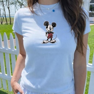 May include: Light blue t-shirt featuring a classic Mickey Mouse graphic. The cartoon character is depicted in black, red, and white. The shirt is a crew neck style and appears to be made of a soft material.