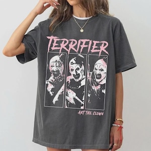 May include: A charcoal gray short-sleeve t-shirt featuring a graphic print of three panels showing a clown character. The text TERRIFIER appears in pink at the top, and ART THE CLOWN is printed in pink at the bottom of the graphic design.