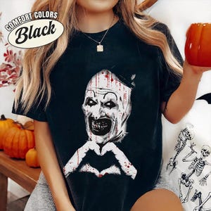 May include: Black t-shirt with a graphic of a scary clown face and a heart-shaped hand gesture. The text "Comfort Colors Black" is in a white oval on the upper left. The shirt is part of a Halloween-themed collection.